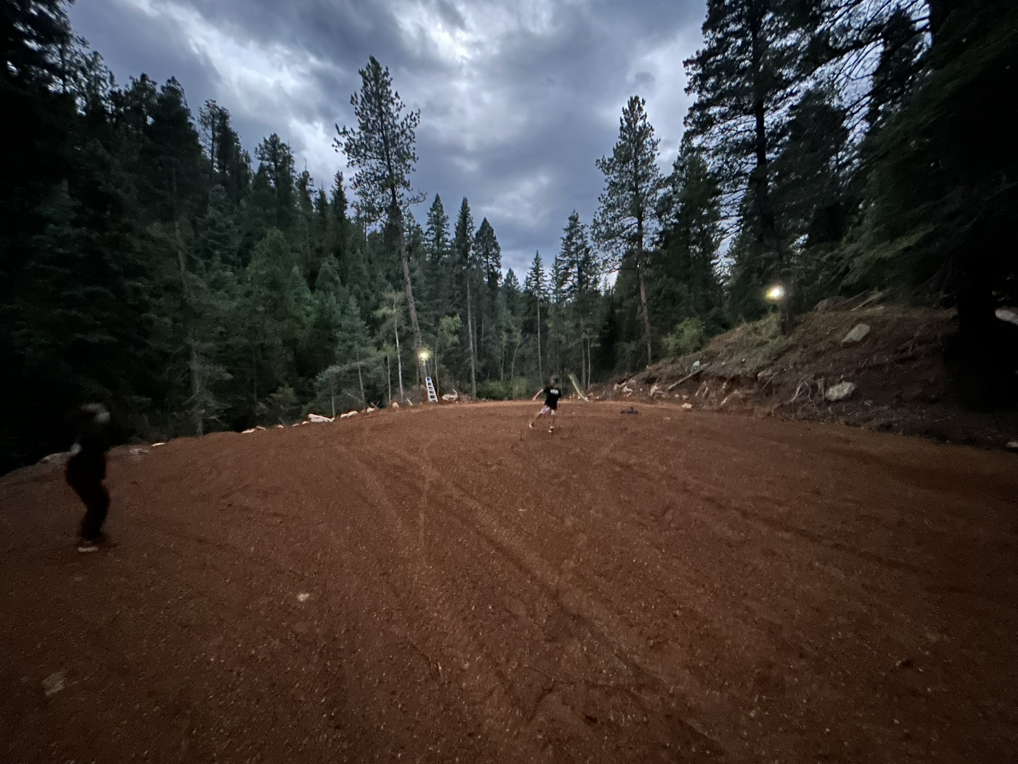 Arena clearing at dusk surrounded by pine forest and lights.